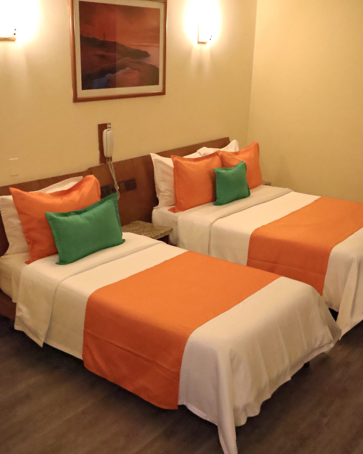 Doble con una cama matrimonial y una individual, cobertores naranja, cojines verdes y cuadro de un atardecer en la costa