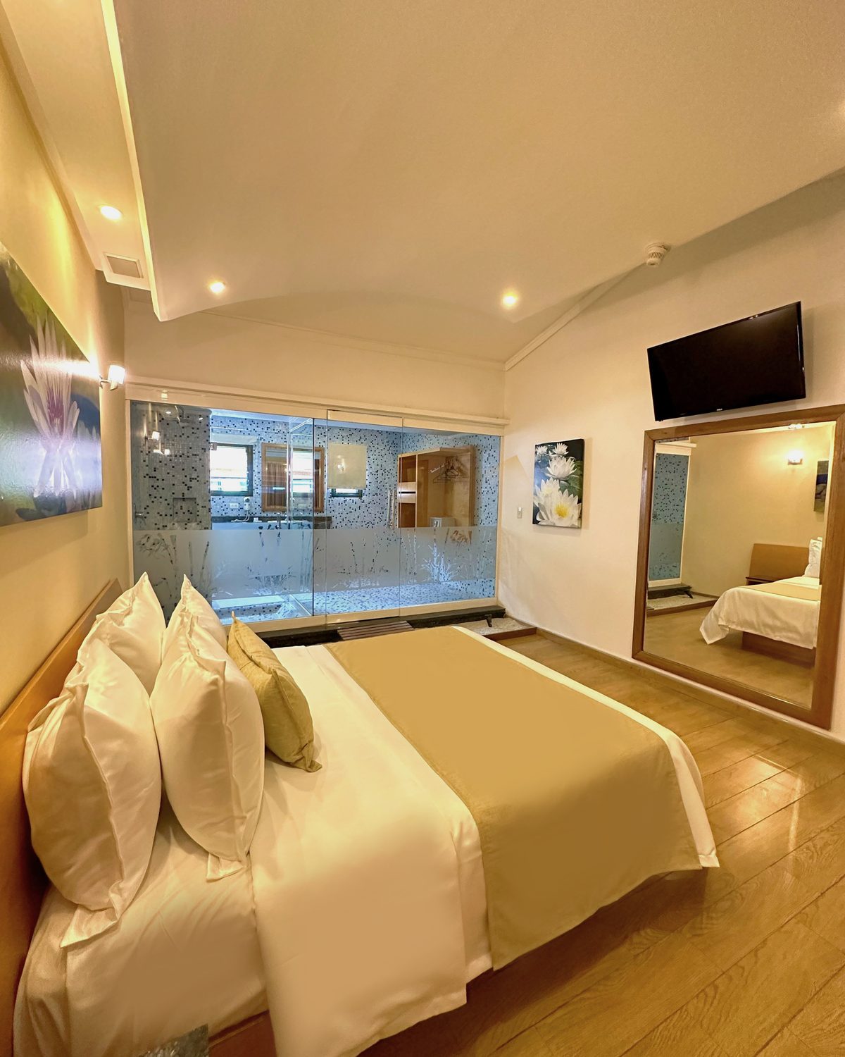 Zen con Terraza: cama con almohadas blancas, televisor, espejo y baño de vidrio al fondo
